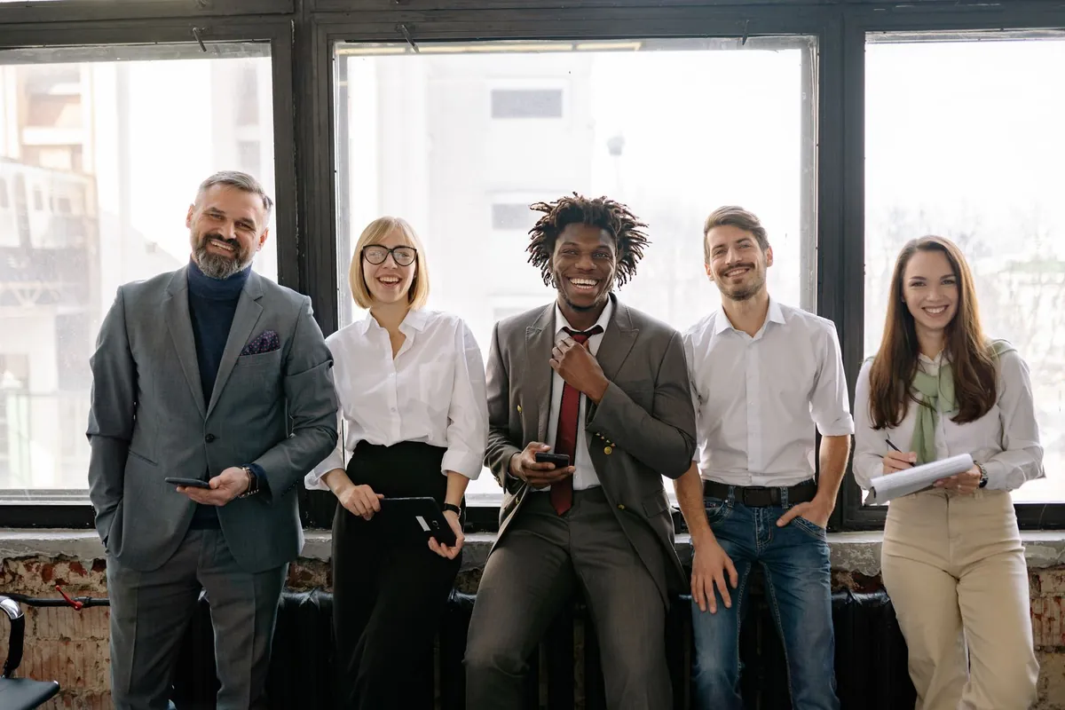 A diverse group of five adults smiling together beside the windows of a bright office