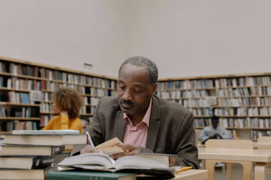 A man reading and studying at a desk in a public library, surrounded by bookshelves